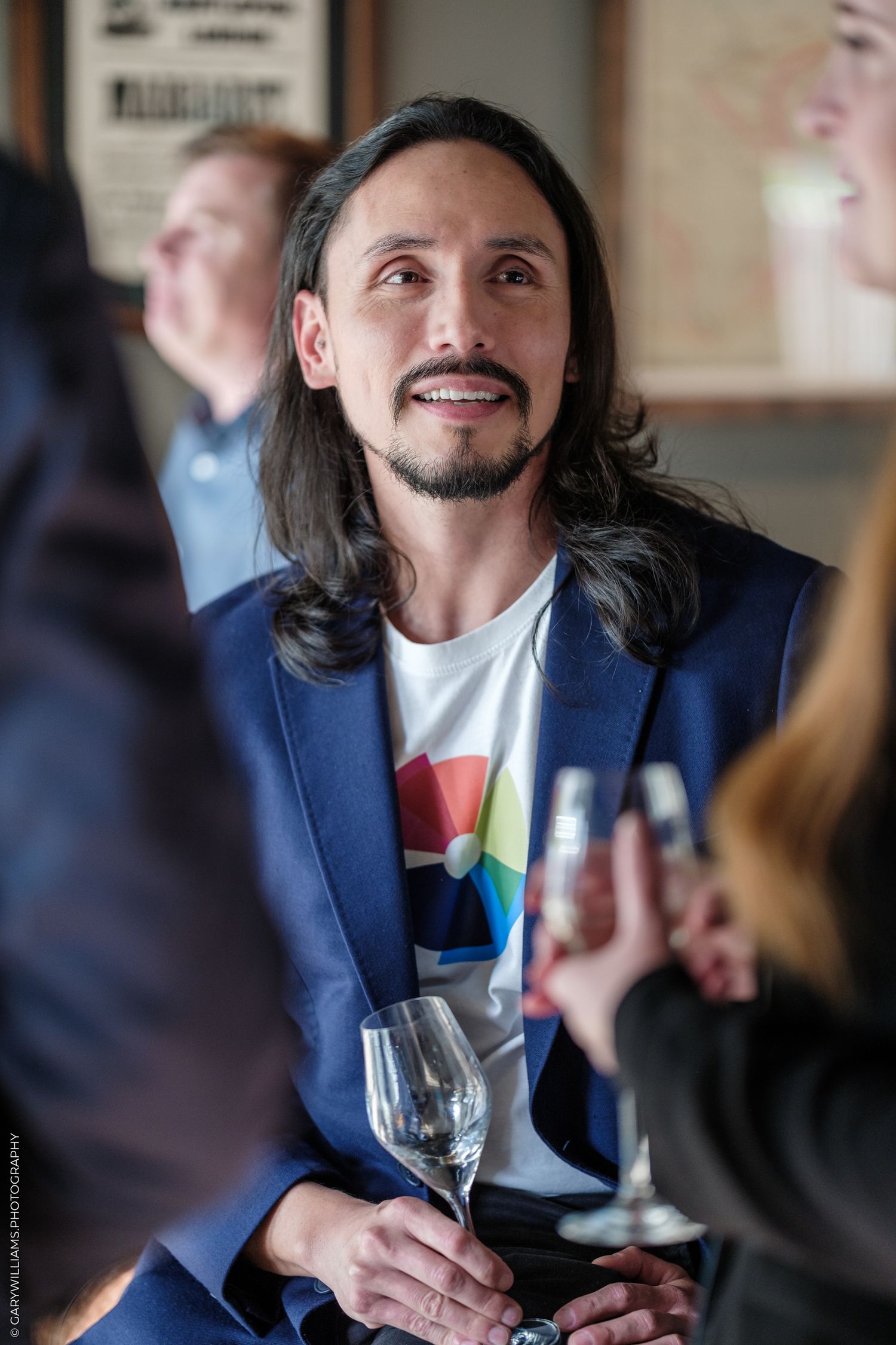 Man with long hair and beard in blue blazer holding wine glass at social gathering.