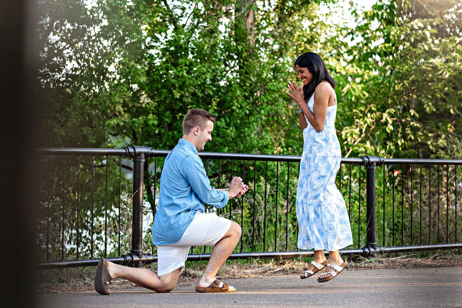 Man kneeling on one knee proposing to a woman in a blue floral dress outdoors near a park railing. Stillwater, MN.  Proposal Photography.