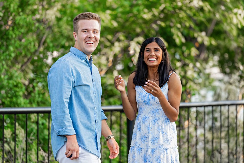Laughing couple standing together outdoors near green trees, man in blue shirt and woman in floral maxi dress.