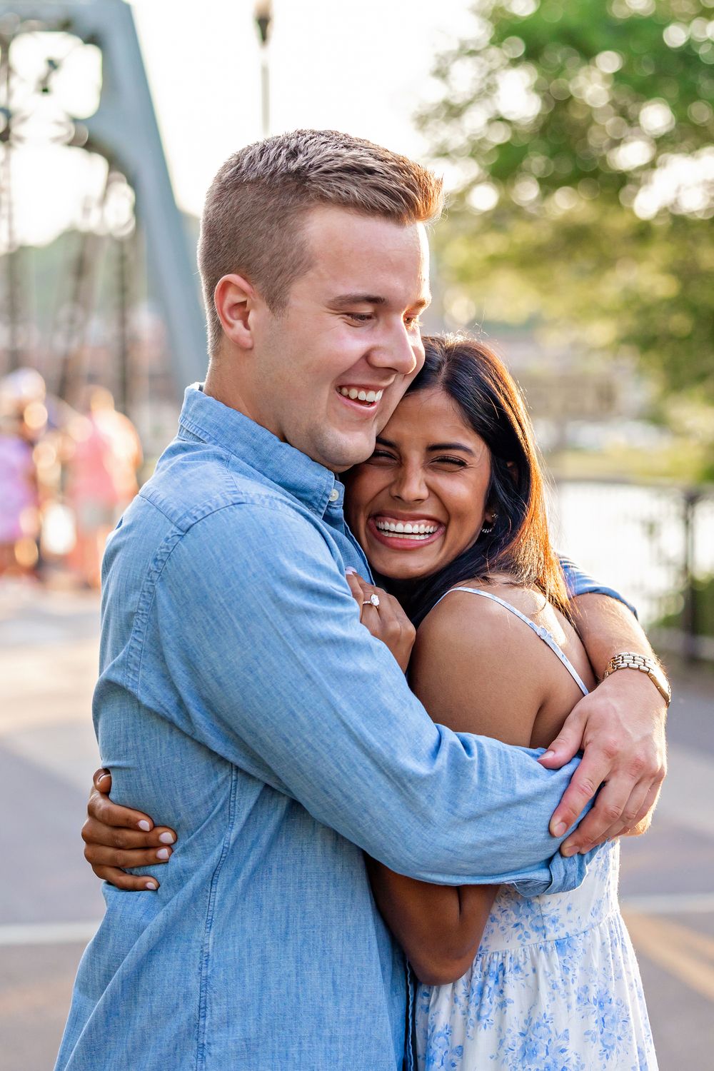 Happy couple laughing and hugging on a bridge, woman in floral dress and man in blue denim shirt.