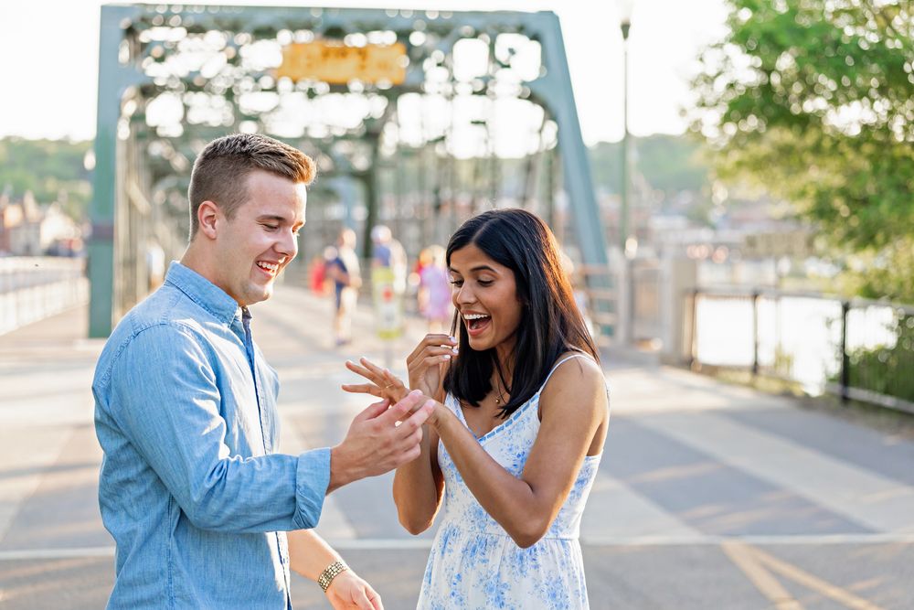 Newly engaged woman admires her ring while her partner smiles on a scenic bridge at golden hour.
