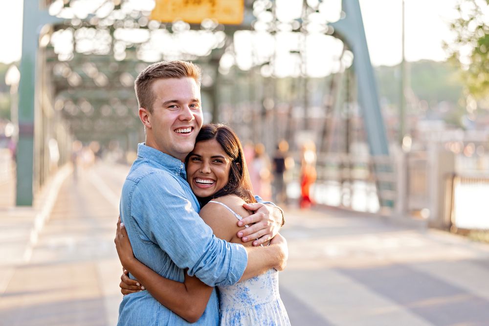 Smiling couple embracing on a bridge, man in blue shirt hugging woman in floral dress at golden hour.