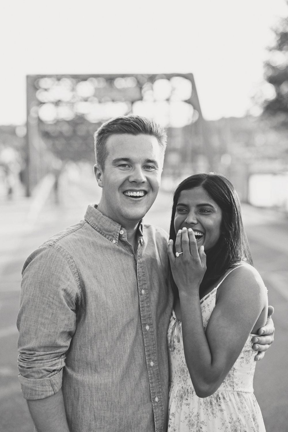Black and white photo of smiling engaged couple, woman showing off engagement ring on a bridge.