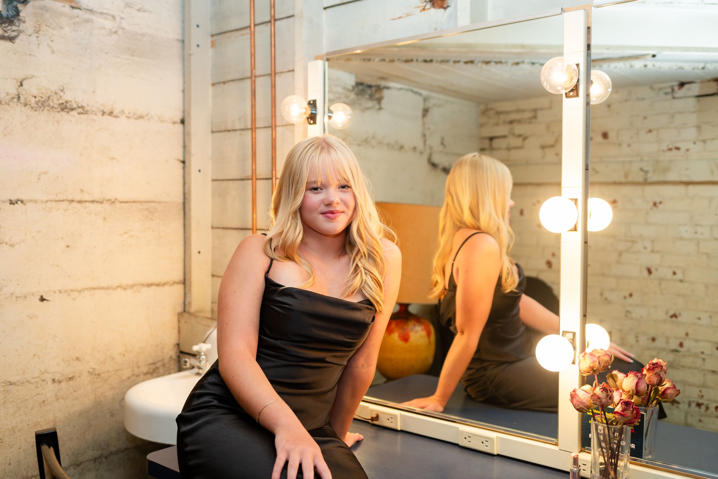 Blonde woman in black dress sits before lit vanity mirror in rustic studio setting.