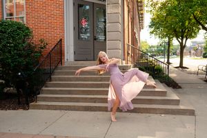 Blonde dancer in pink dress performing graceful pose on sidewalk outside brick building with steps.