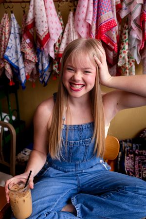 Happy blonde girl in denim overalls laughing while holding an iced coffee in a colorful cafe.