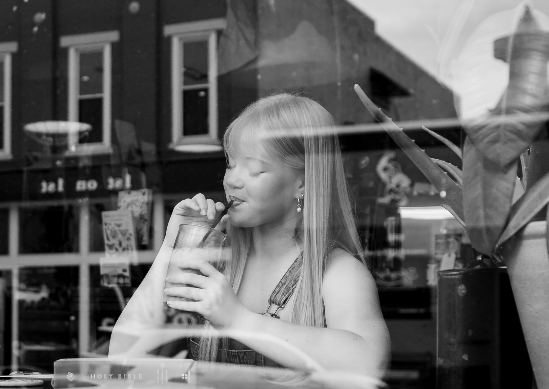 Black and white photo of a young woman sipping a drink through a straw at a cafe window seat.