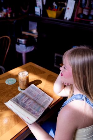 Young woman reading a book at a wooden cafe table with an iced coffee drink in warm sunlight.