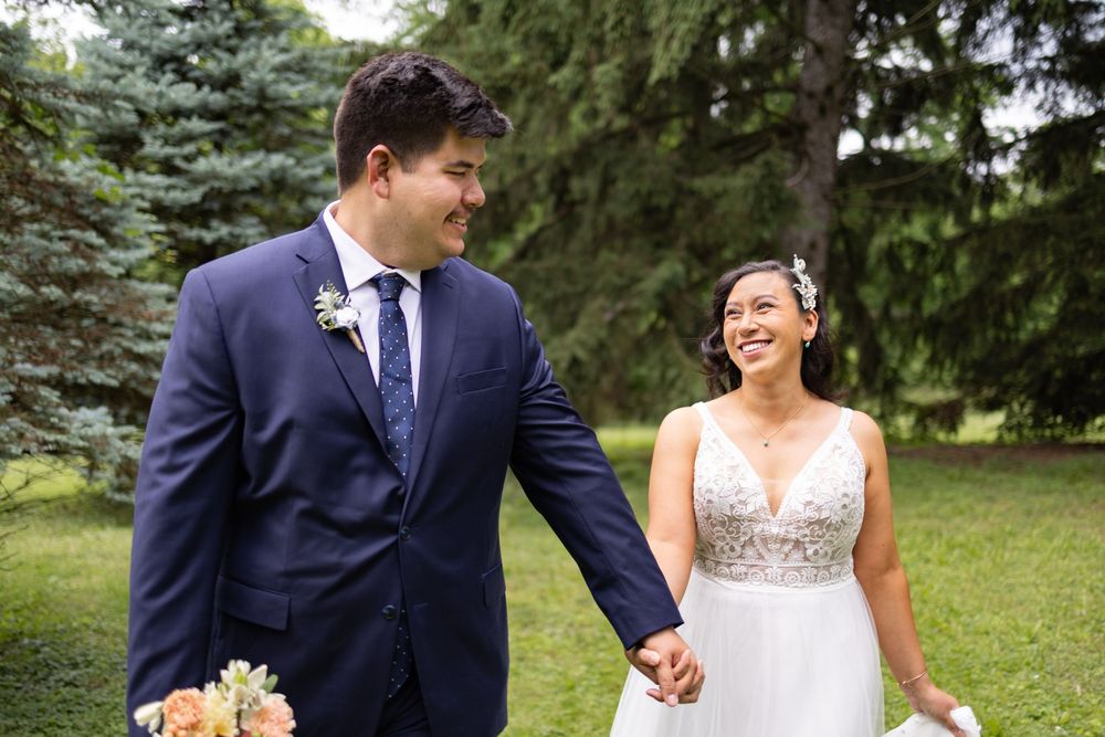 A smiling bride in a white lace gown holds hands with a groom in a navy suit outdoors among green trees.