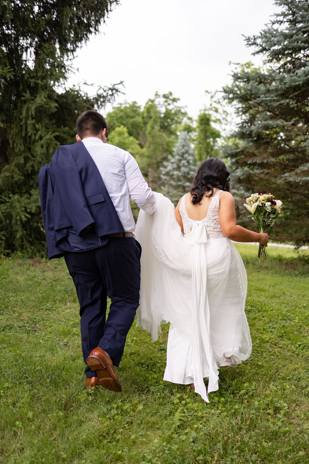 Bride and groom walking away through lush green garden, bride in white lace gown holding bouquet, groom in navy suit.