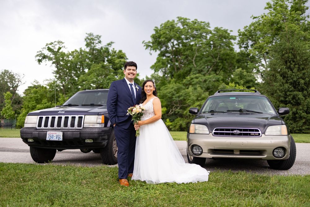 Bride and groom posing between two SUVs outdoors on their wedding day, bride holding a bouquet.