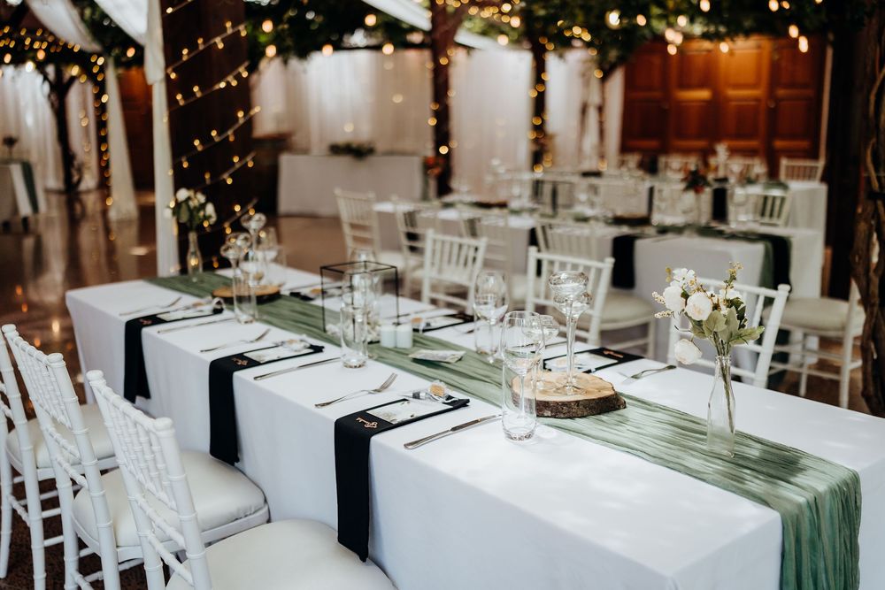 Elegant wedding reception table with white linens, sage green runner, black napkins, and fairy lights in a rustic venue.