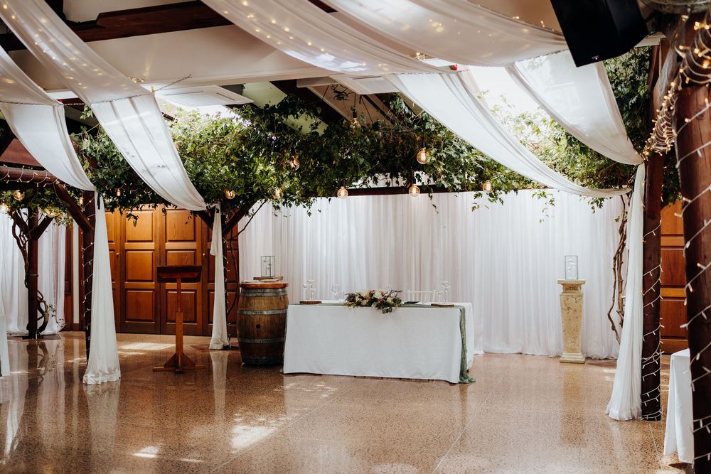 Elegant wedding reception venue with white draping, fairy lights, lush greenery ceiling, and a decorated sweetheart table.
