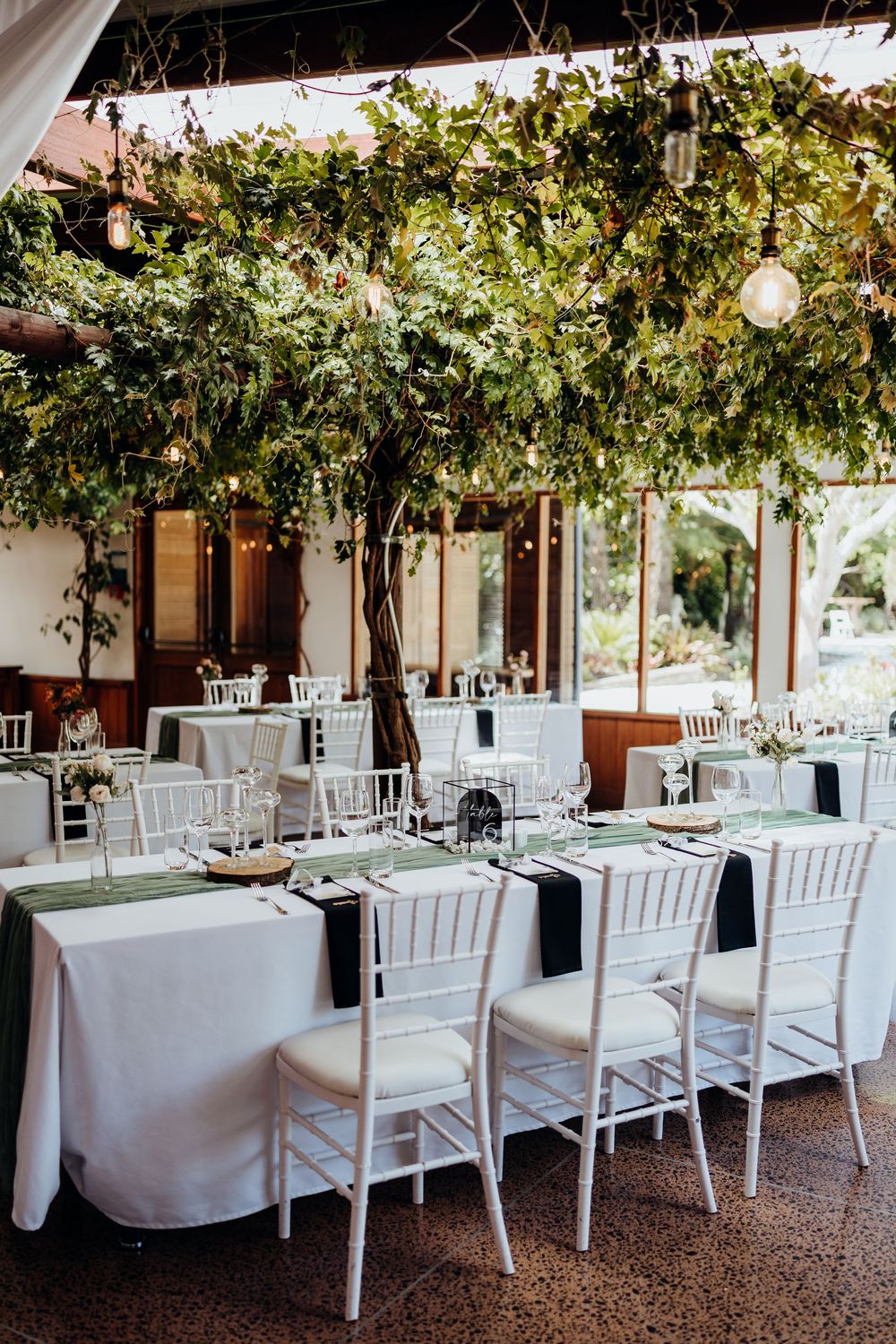 Elegant wedding reception with white Chiavari chairs, green table runners, and lush greenery overhead with string lights.