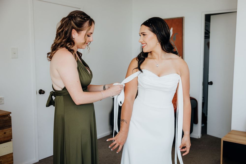 Bridesmaid in olive green dress helping bride with white wedding gown bow tie during bridal preparations.