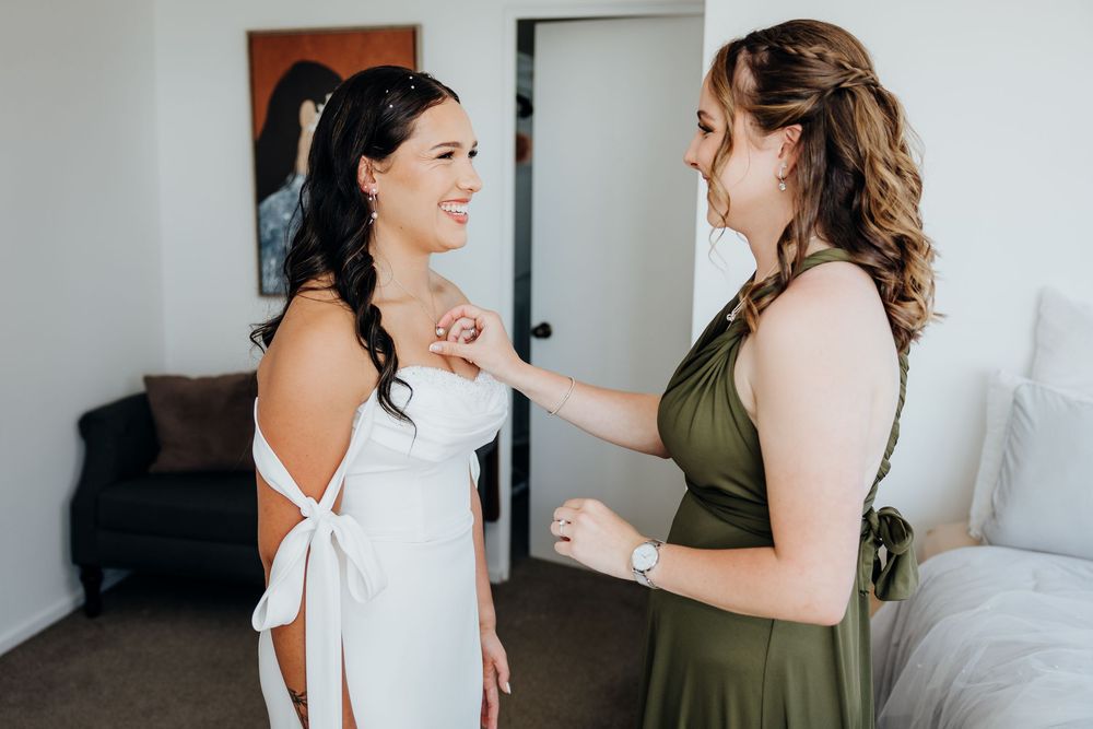 Bride in white backless gown smiling with bridesmaid in olive green dress during wedding preparations.