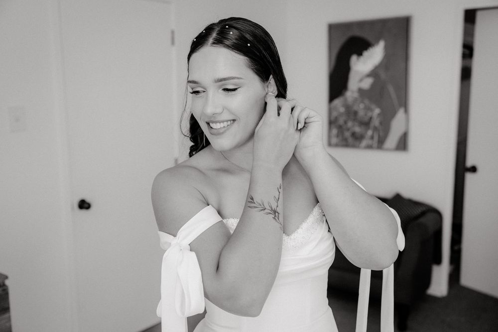 Bride in white off-shoulder gown putting on earrings in black and white photo.