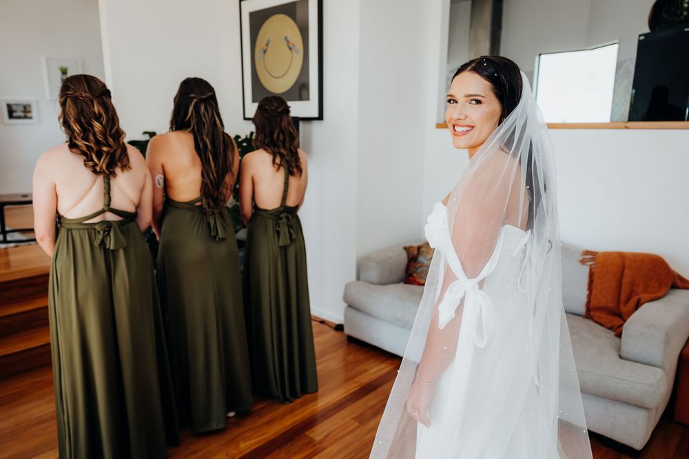 Bride in white wedding gown and veil smiles over shoulder while bridesmaids in olive green dresses stand nearby.