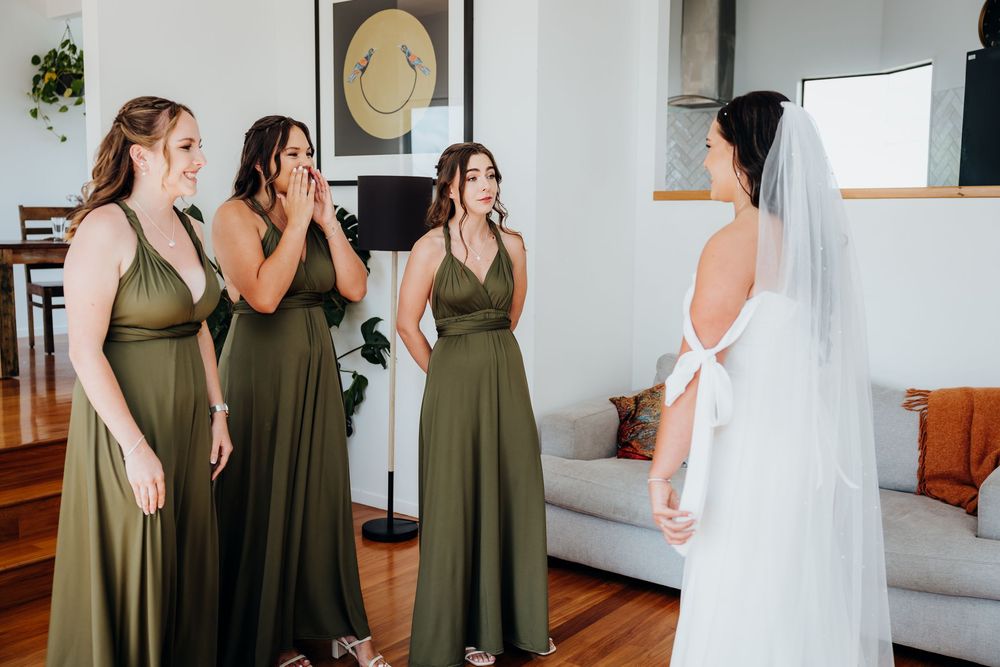 Three bridesmaids in olive green dresses reacting with joy seeing bride in white wedding gown and veil.