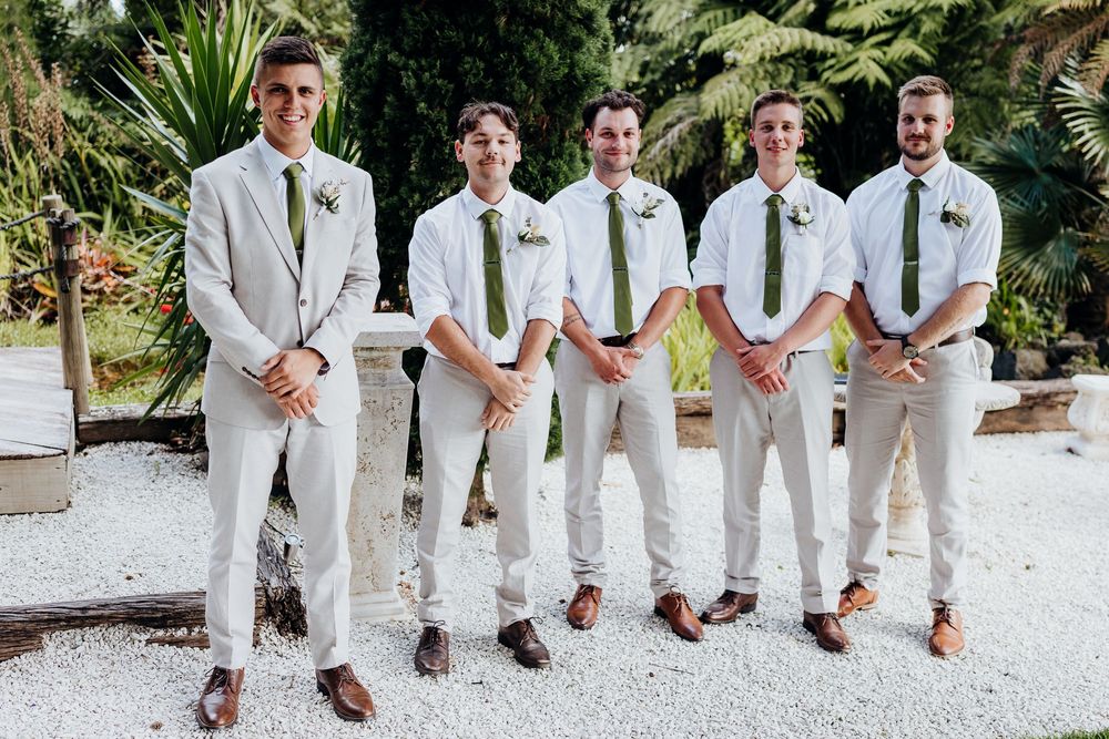 Five groomsmen in matching tan suits with green ties and brown dress shoes pose together outdoors at a wedding.