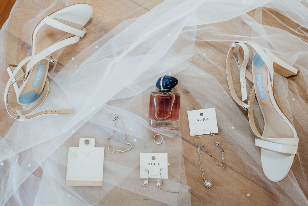 Bridal flat lay featuring white heeled sandals, perfume bottle, AIRA earrings, and a delicate veil on wooden floor.