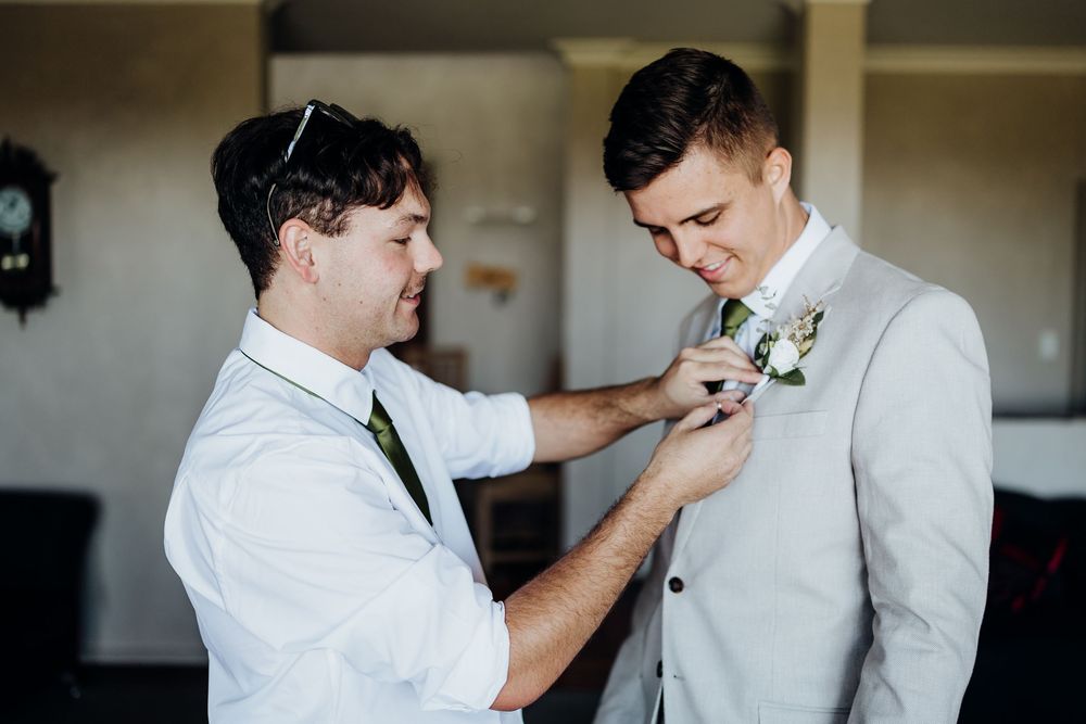 Groomsman pinning a white boutonniere on groom in light gray suit before wedding ceremony.