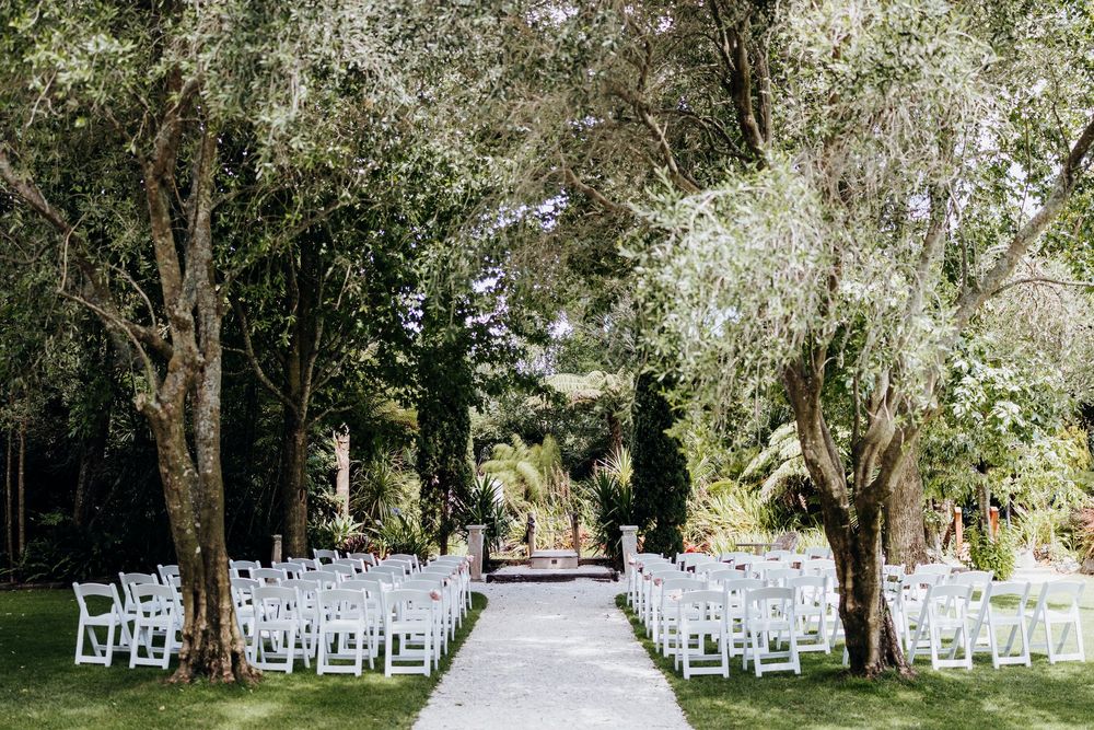 Elegant outdoor wedding ceremony setup with white chairs lining a tree-lined aisle in a lush garden.