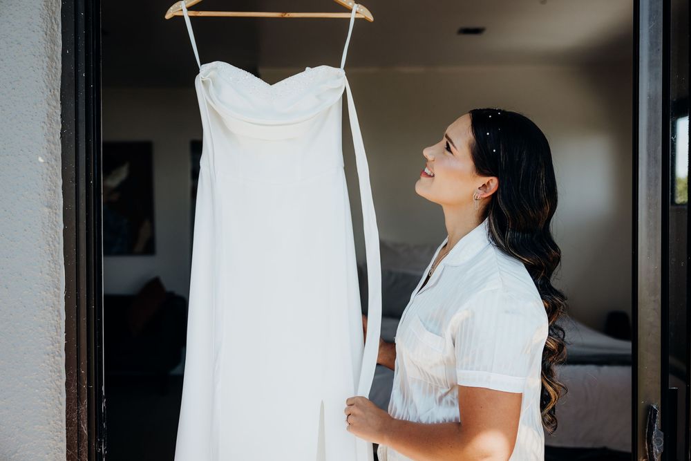 Bride admiring her white wedding gown hanging in doorway during bridal preparations.
