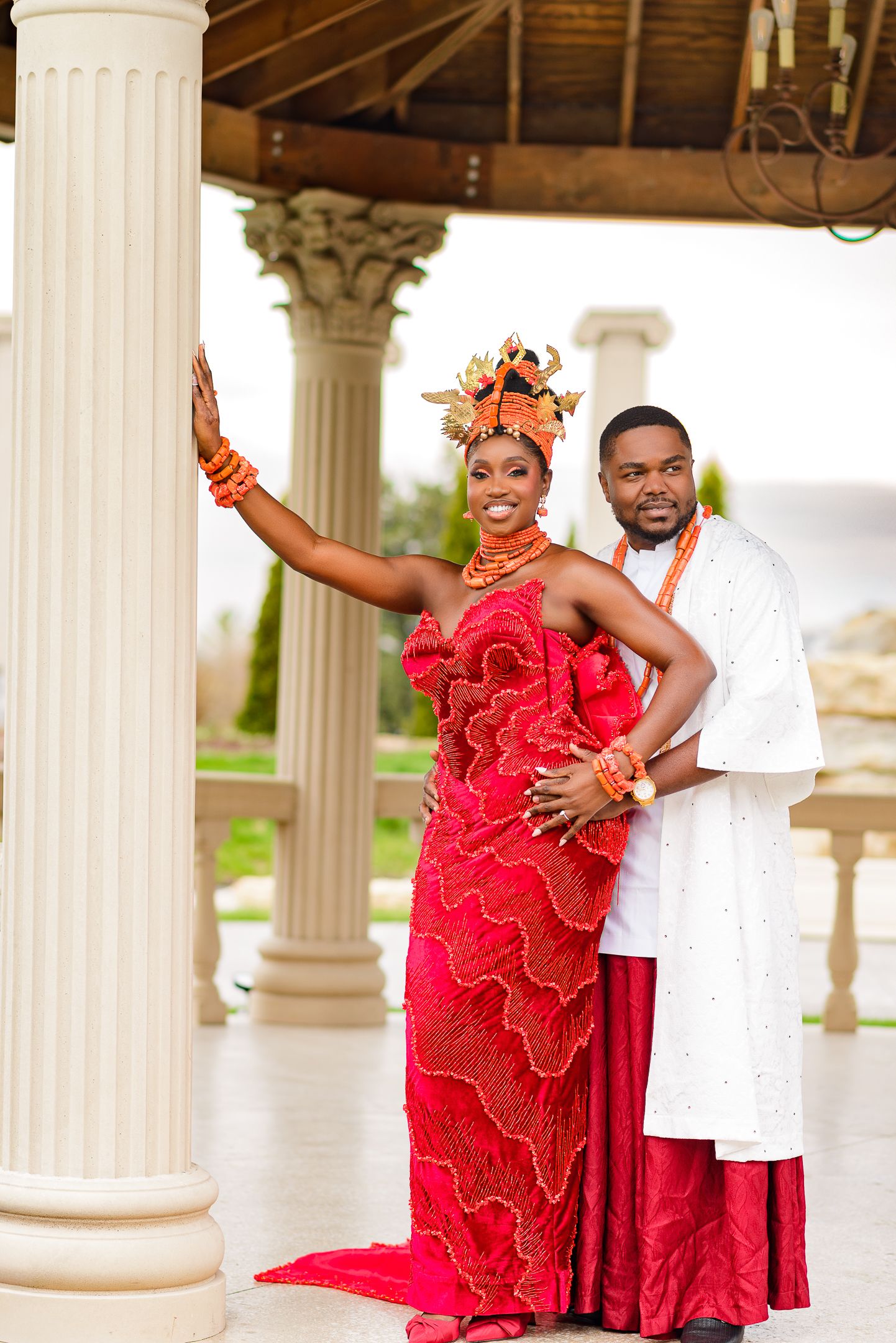 Couple in traditional Nigerian attire, woman in red gown with headdress, man in white agbada, posing under a gazebo.