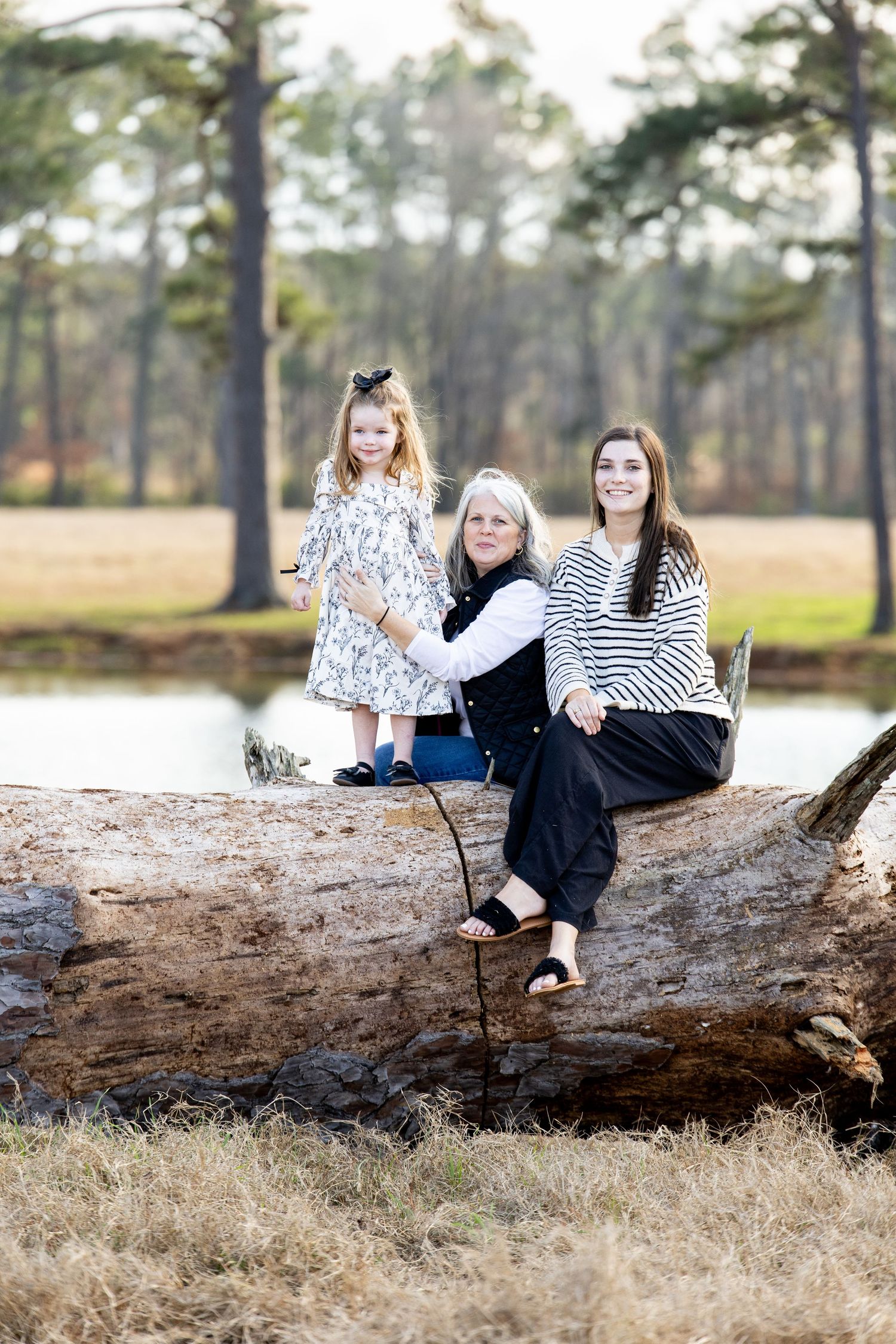 Three generations of women pose on a fallen log near a pond surrounded by tall pine trees.