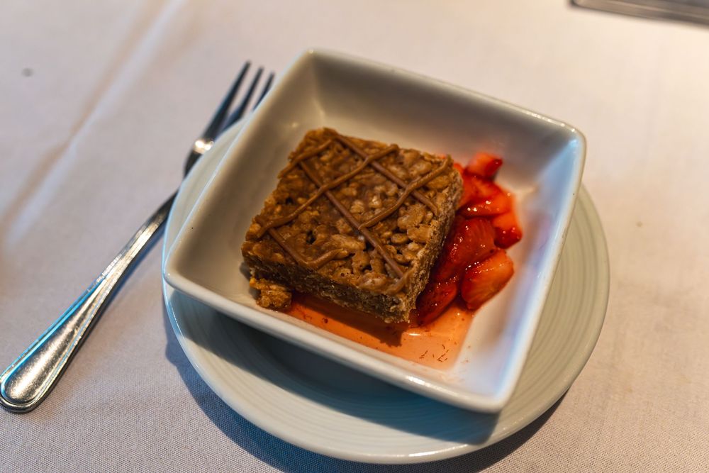 Square white dish with layered chocolate brownie dessert topped with nuts and fresh strawberries on a restaurant table.