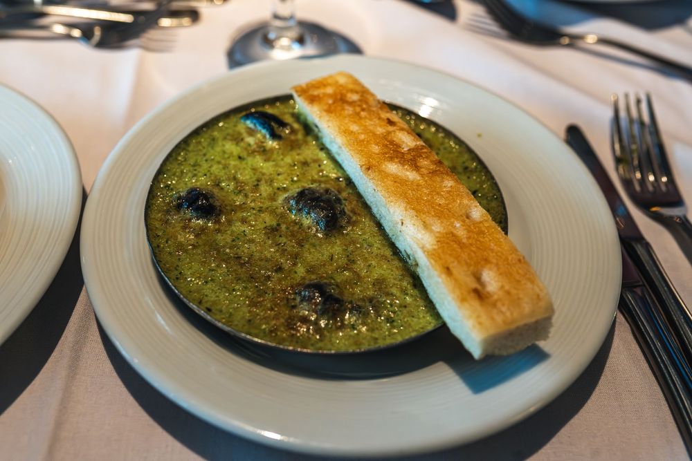 Elegant restaurant dish featuring green herb sauce with escargot and a slice of toasted bread on a white ceramic plate.