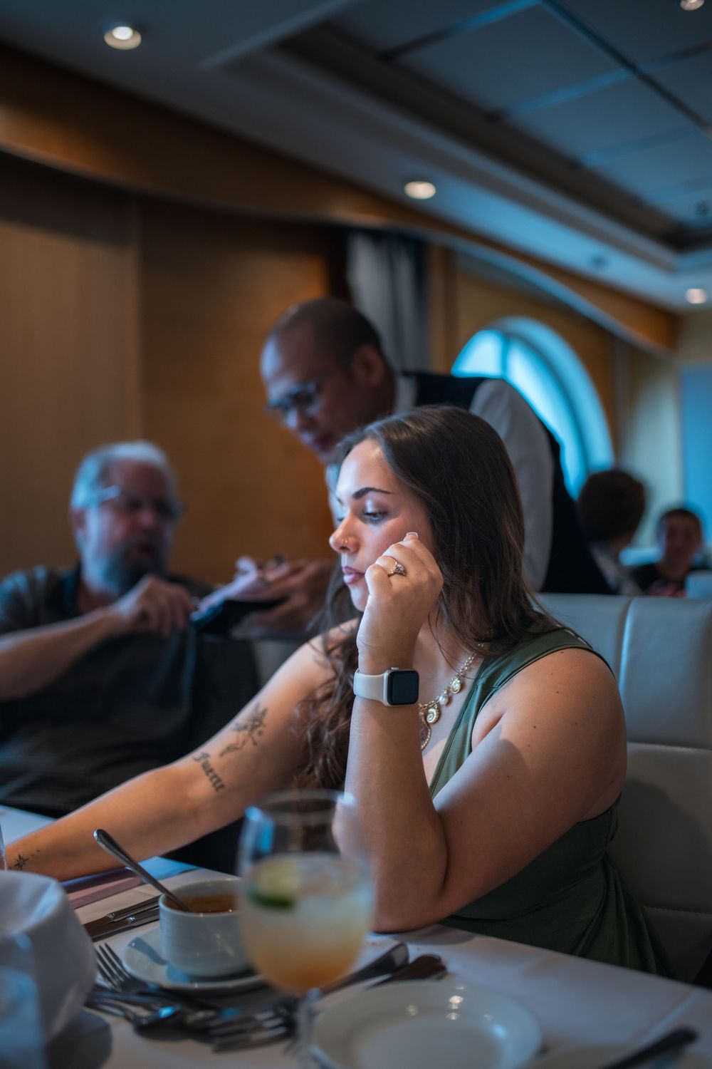 Woman with a tattoo and a smartwatch looks at her phone while dining in an upscale restaurant with others nearby.