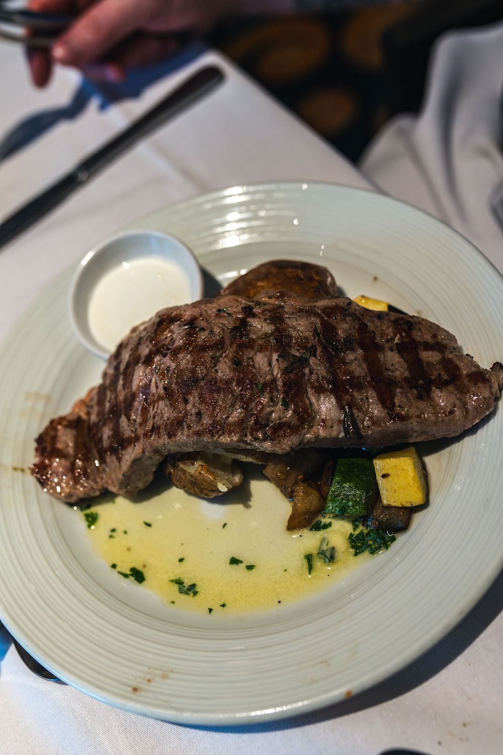 Grilled ribeye steak served with roasted vegetables, herb butter sauce, and a side of creamy dipping sauce on a white plate.