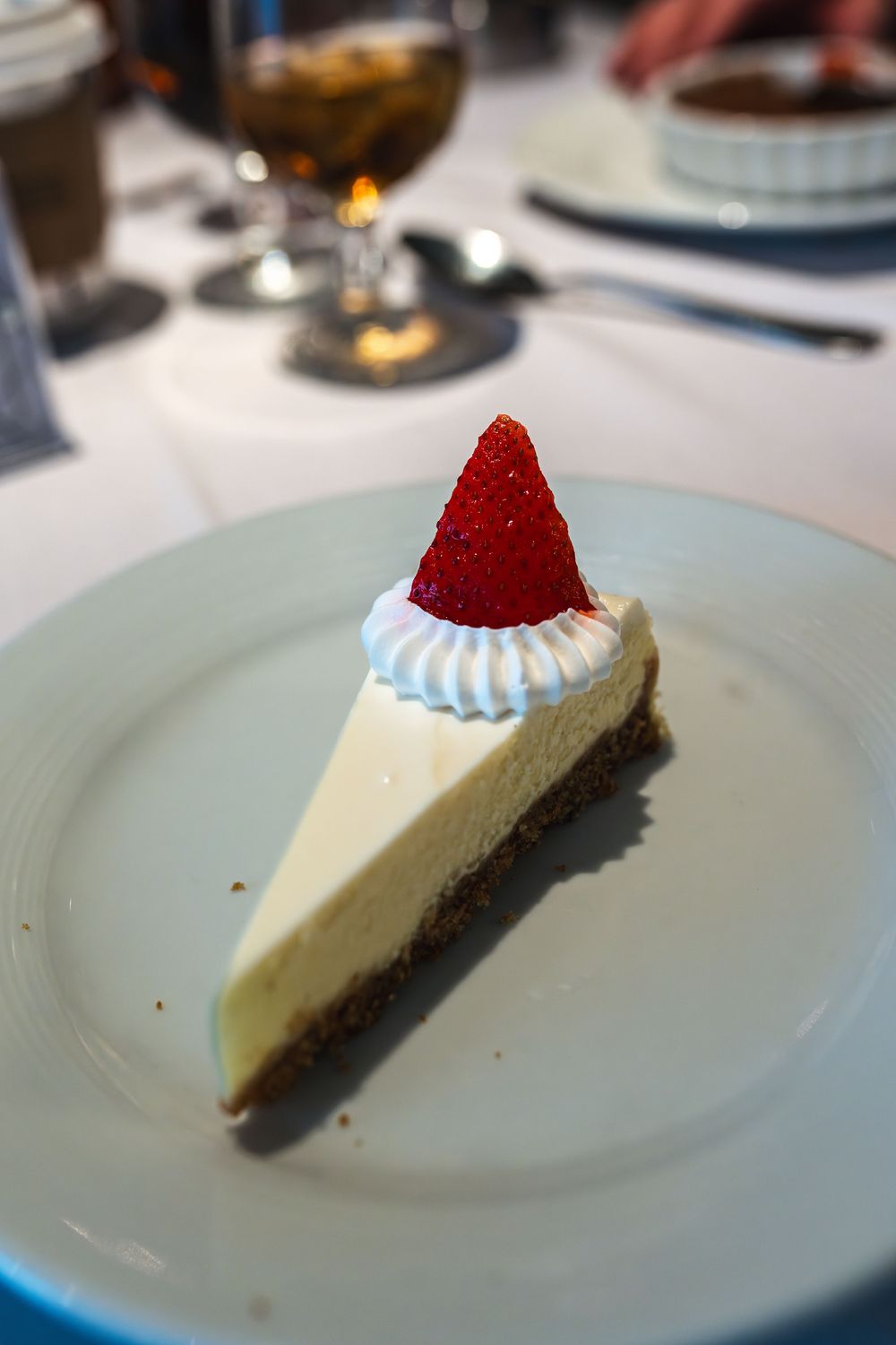 A slice of creamy New York cheesecake topped with whipped cream and a fresh strawberry on a white plate.