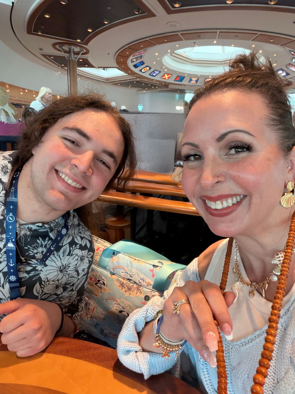 Two smiling people pose for a selfie inside a cruise ship atrium, one wearing a floral shirt and lanyard.
