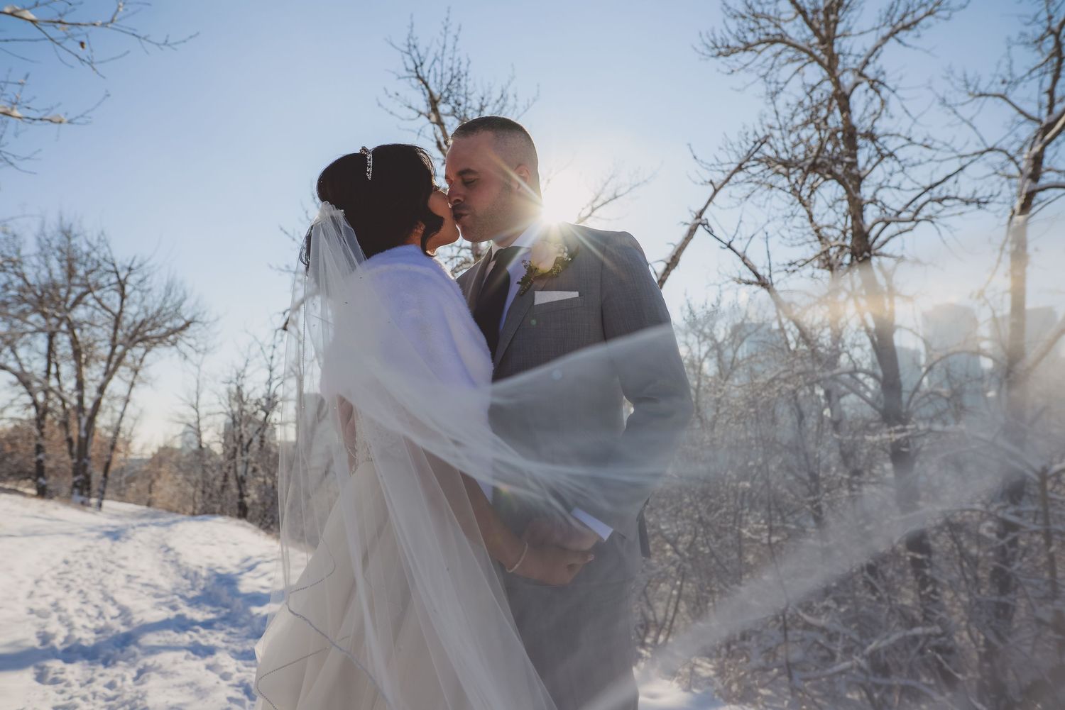 Bride and groom share a kiss outdoors in a snowy winter landscape, with sunlight and bare trees in the background.