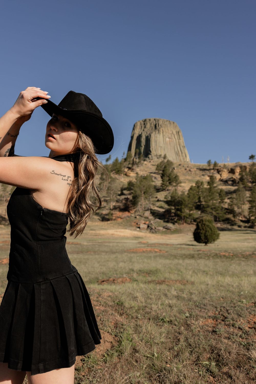 Woman in black cowboy hat and mini dress poses outdoors near Devils Tower, Wyoming under clear blue sky.