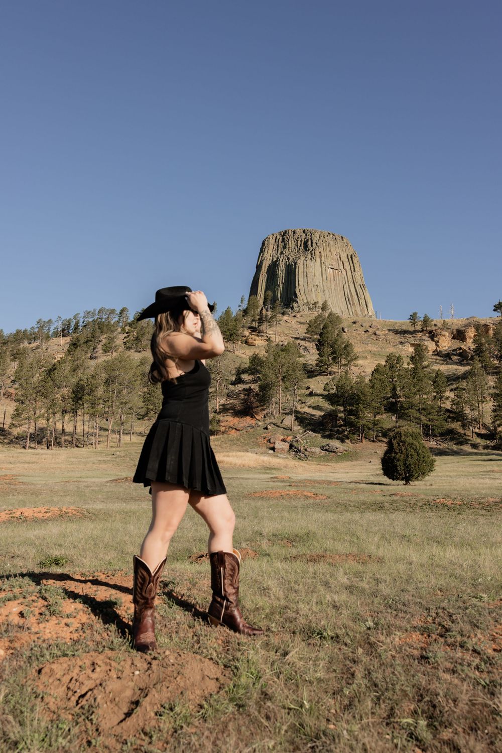 Woman in black dress, cowboy hat, and boots poses near Devils Tower, Wyoming under a clear blue sky.