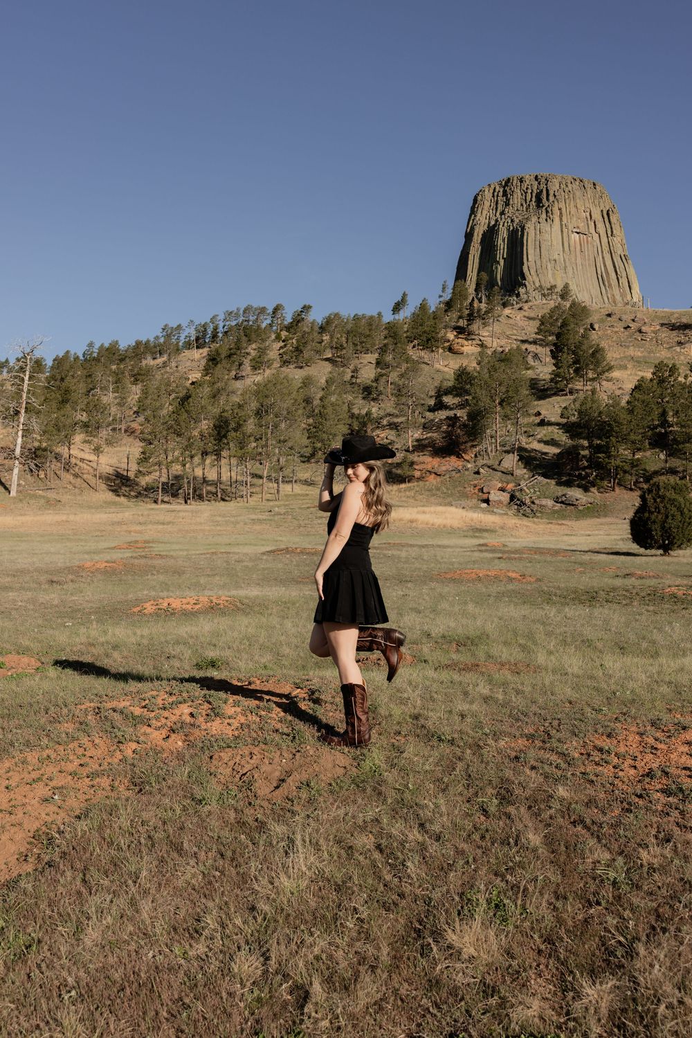 Woman in black dress and cowboy hat poses near Devils Tower, Wyoming, surrounded by pine trees and red soil.