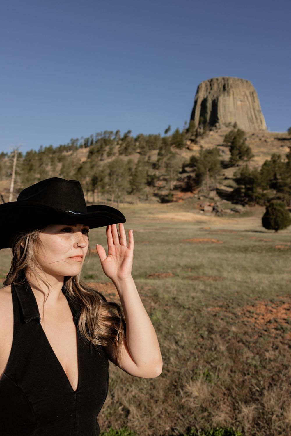 Woman in black cowboy hat shielding eyes near Devils Tower, Wyoming landmark rising above green meadow.