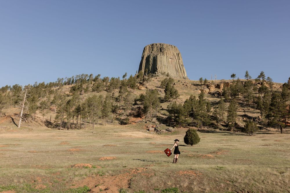 Person walking near Devils Tower National Monument, Wyoming, with rocky butte and pine trees under clear blue sky.