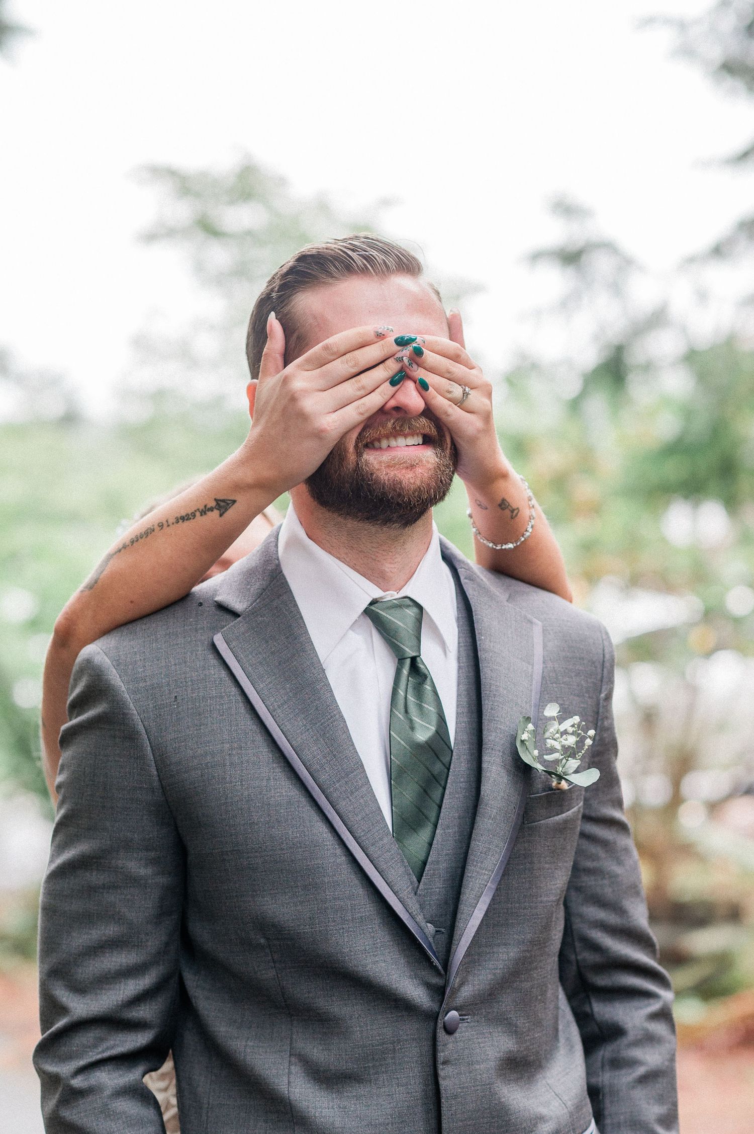 Groom in gray suit with green tie has eyes covered by bride's hands during wedding first look.