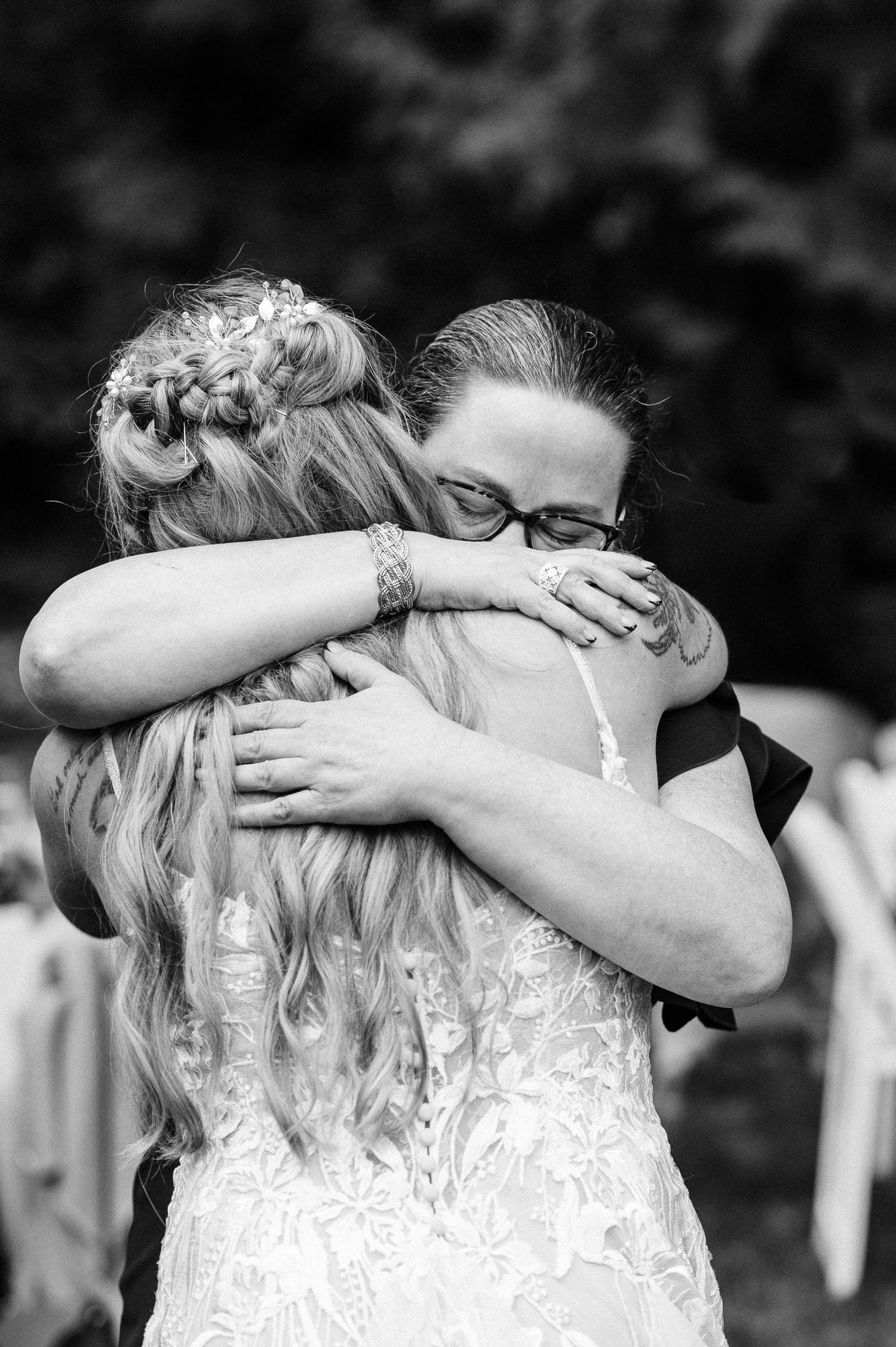 Black and white photo of a bride in lace dress embracing mother in glasses at an outdoor wedding ceremony.