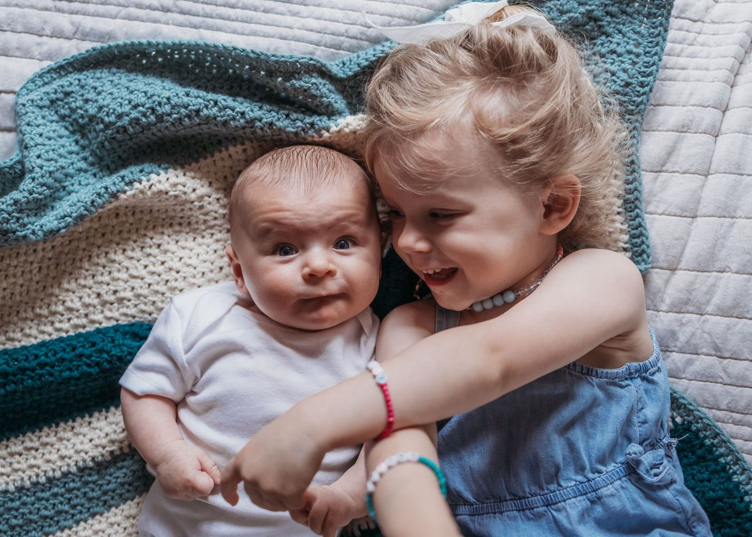A smiling toddler girl hugs a newborn baby sibling while lying on a knit blanket.