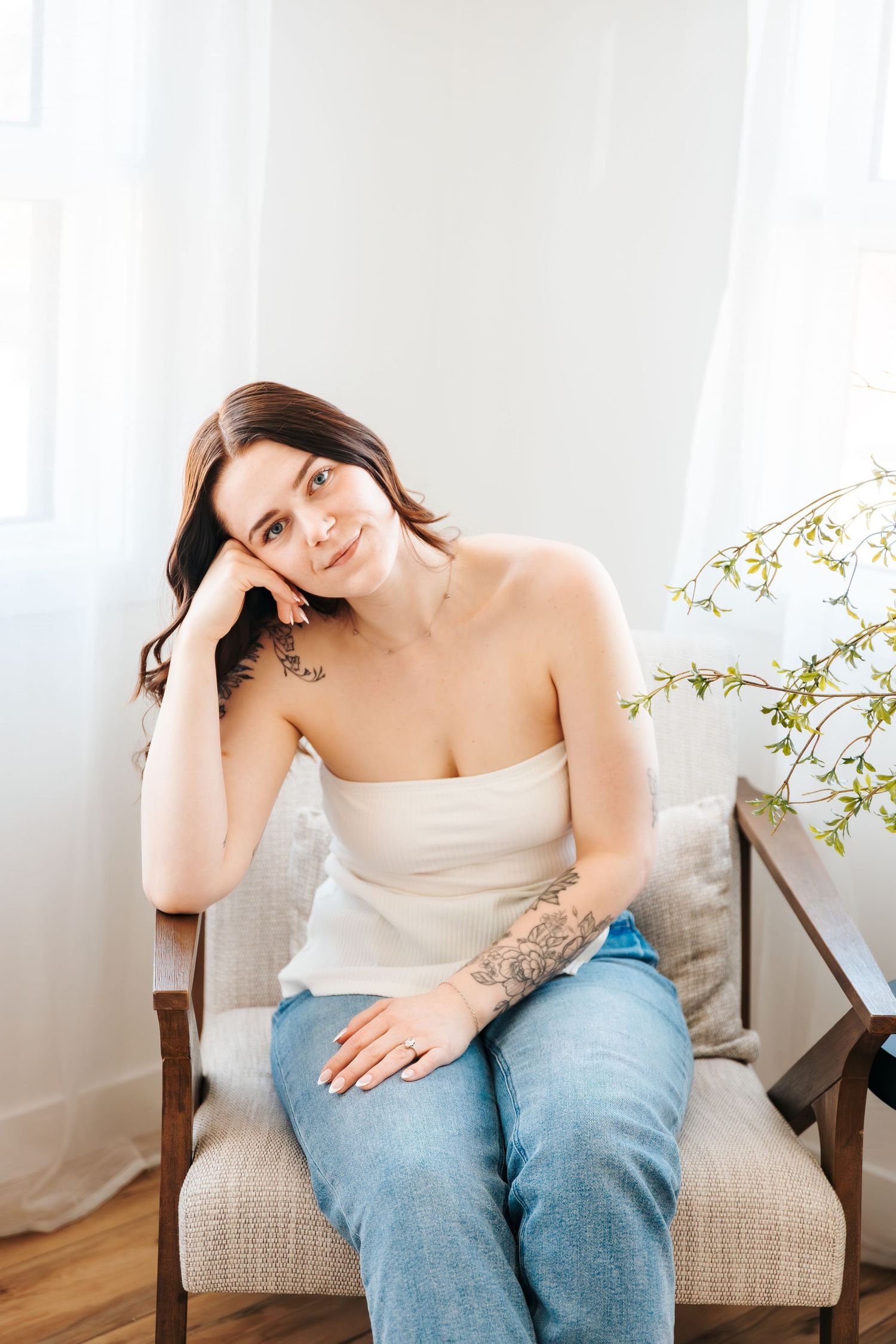 A woman with tattoos in white top and jeans sitting relaxed in armchair near bright window. Edmonton area photographer. Business photos