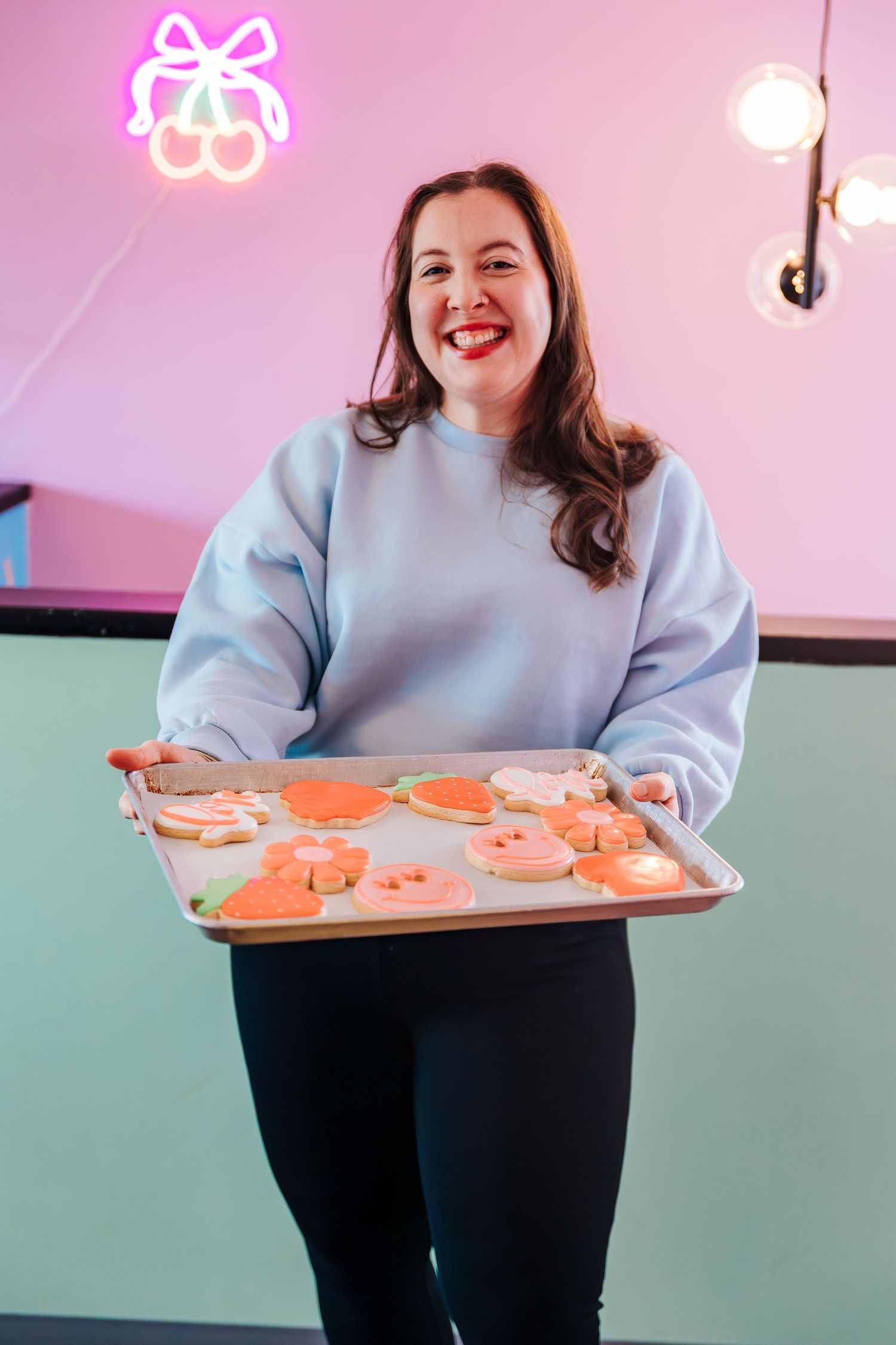 Smiling woman in blue sweatshirt holding a tray of colorful decorated cookies in a pink bakery. Headshots for bakery. Cookies and cakes