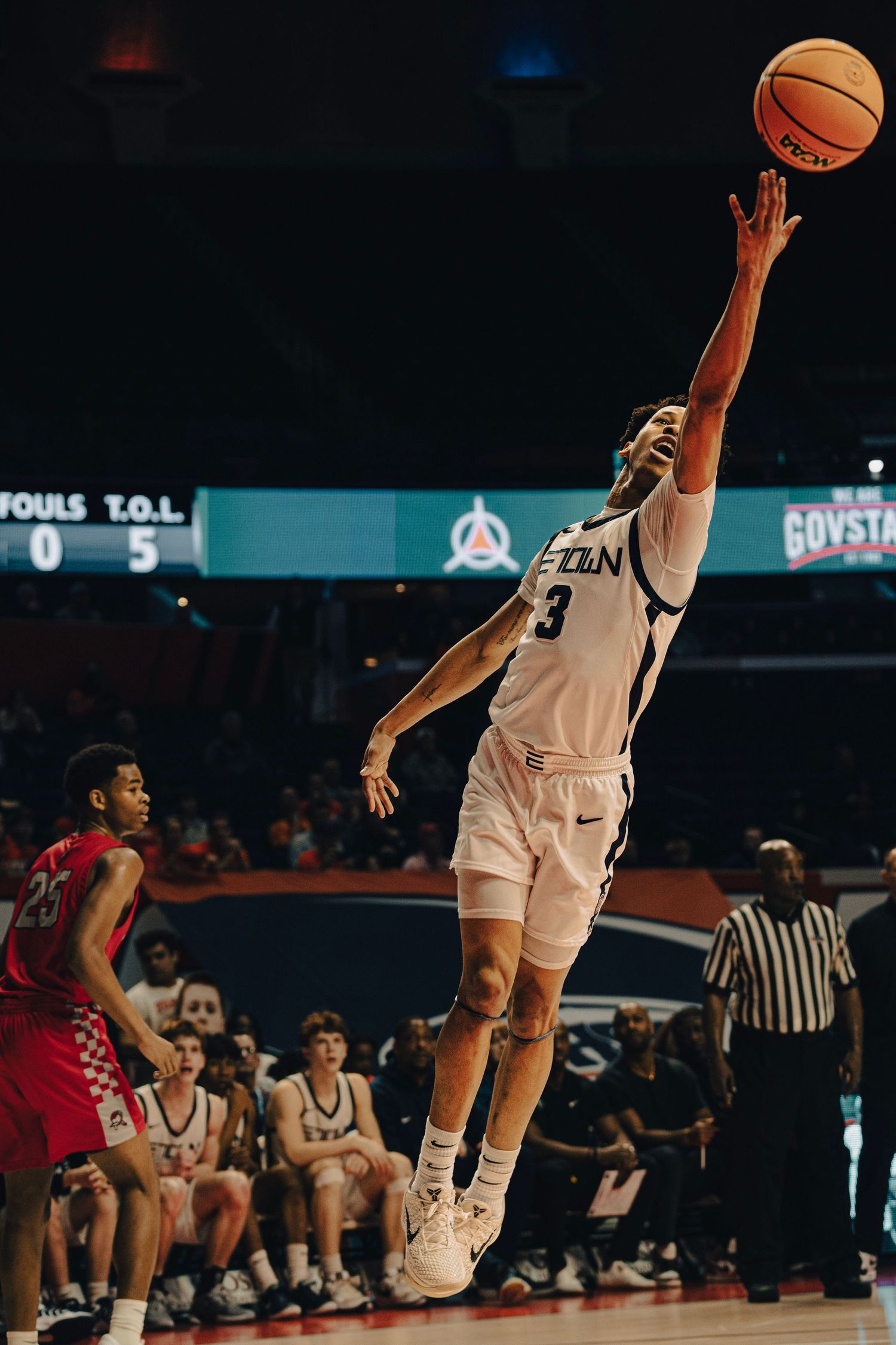 Basketball player #3 in white uniform leaps to shoot during an indoor college basketball game under bright arena lights.