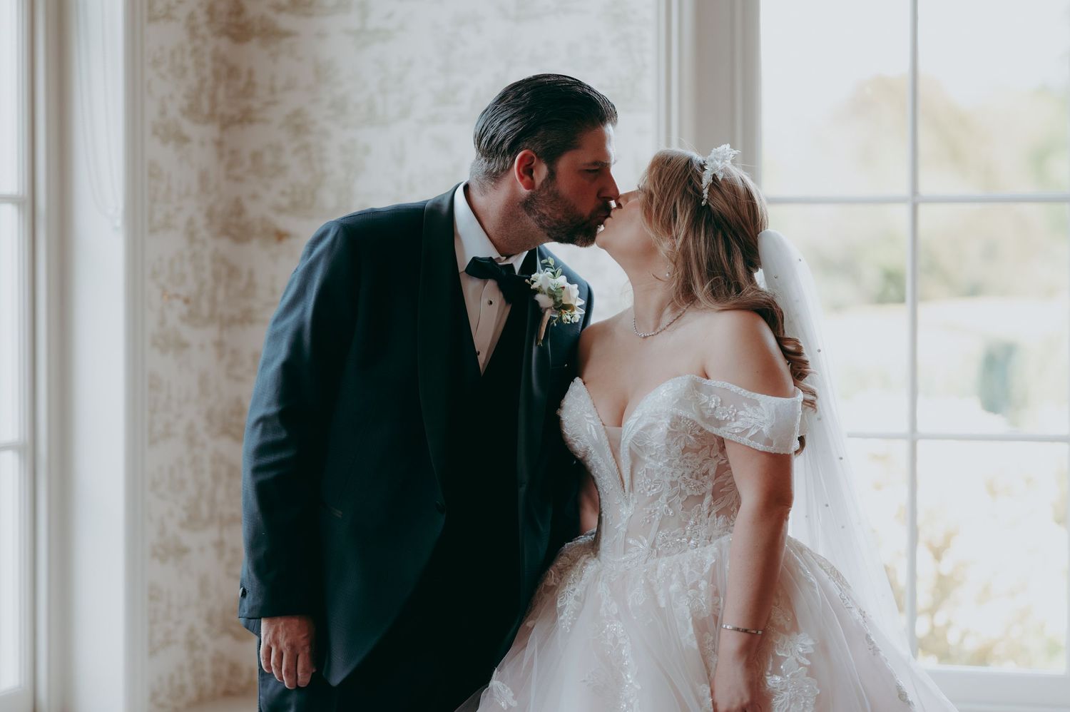 Bride and groom sharing a romantic kiss near a window, bride in off-shoulder lace gown with veil, groom in navy suit.