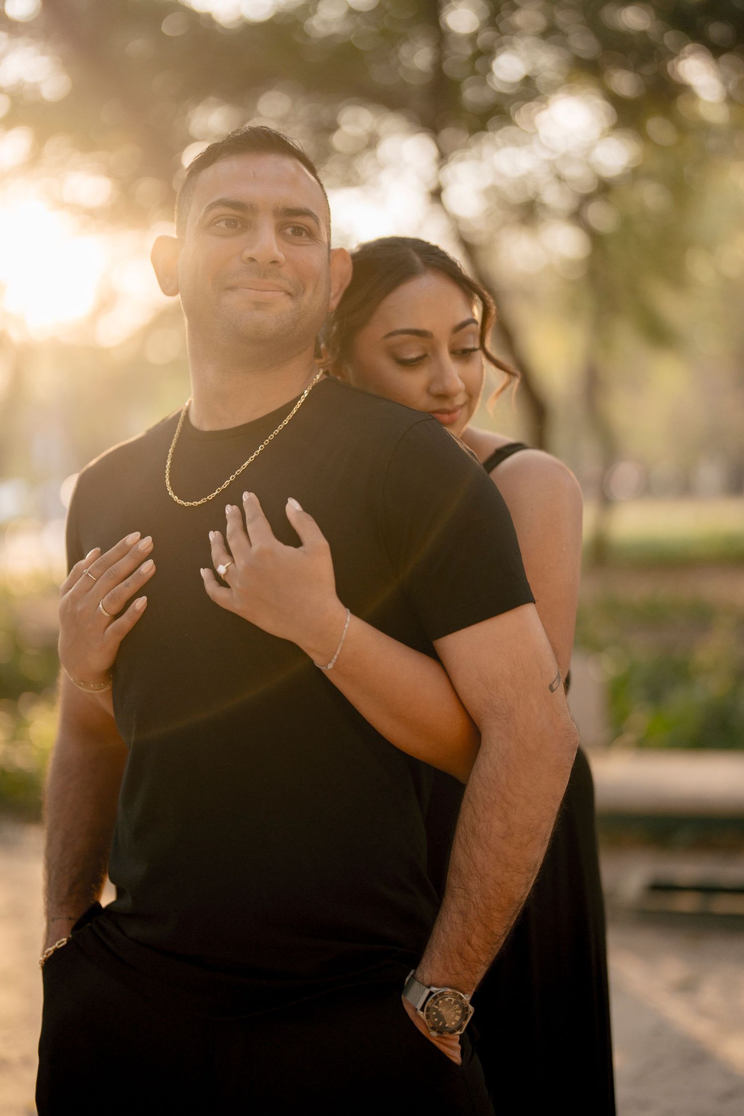 Couple in matching black outfits embracing at golden hour in a park, woman hugging man from behind.